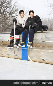 Two boys in ice hockey uniforms sitting on ice rink sidelines looking and smiling.