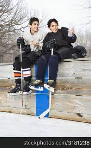 Two boys in ice hockey uniforms sitting on ice rink sidelines pointing and looking.