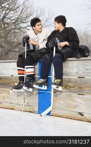 Two boys in ice hockey uniforms sitting on ice rink sidelines talking to eachother.