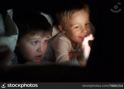 Two boys lying on bed under blanket at night. They watching movie or cartoon on pad. Screen enlighting their faces in darkness