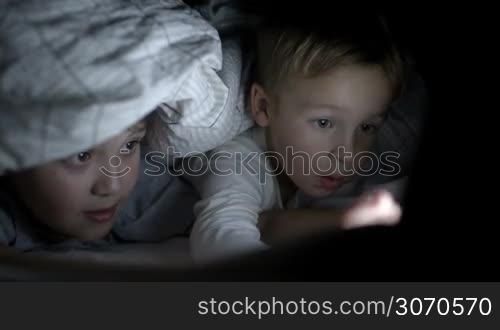 Two boys lying under blanket in bed at night watching cartoon or movie on touch pad
