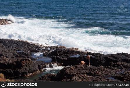 Two boys on rugged coast looking to dive into small pool by raging sea