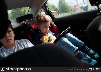 Two boys on the back seat of a car. Elder boy looking on the road, younger one using pad sitting in child safety seat