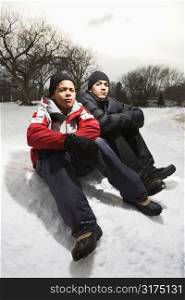 Two boys sitting in snow wearing coats and hats.