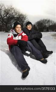 Two boys sitting in snow wearing coats and hats and smiling.