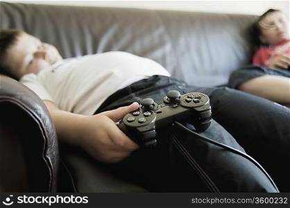 Two boys sleep on sofa holding games consoles