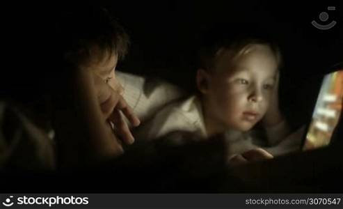 Two boys using touch pad lying in bed at night. Bright screen enlighting their faces