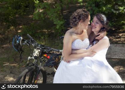 two brides have fun on bench in forest with mountain bikes