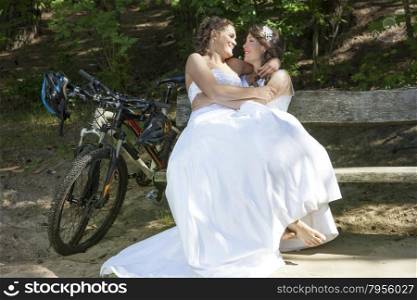 two brides have fun on bench in forest with mountain bikes