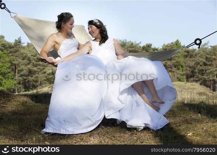 two brides look at each other in hammock against blue sky with forest background