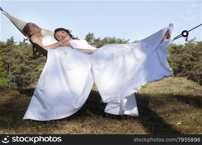 two brides relax in hammock against blue sky with forest background