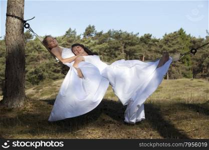 two brides relax in hammock against blue sky with forest background