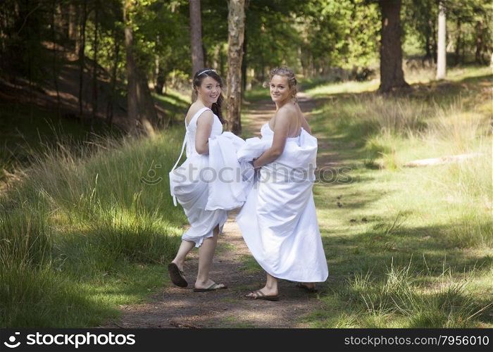 two brides walk on forest path with skirts in their arms while looking back smiling