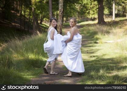 two brides walk on forest path with skirts in their arms while looking back smiling