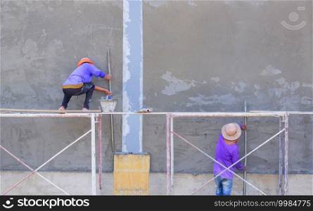 Two builder workers using long trowels to plastering cement on concrete wall of industrial building structure in construction site, rear view with copy space