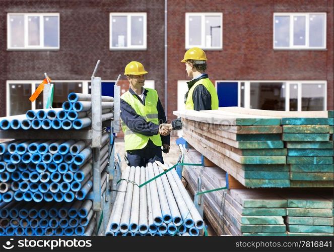 Two building contractors shaking hands behind stacks of scaffolding material, in front of a newly completed residential building complex