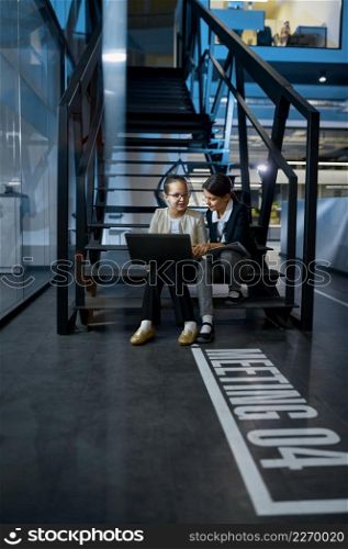 Two business children with laptop and paper document working together sitting on stairs in the office. Two business children working together on stairs