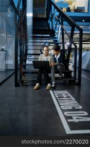 Two business children with laptop and paper document working together sitting on stairs in the office. Two business children working together on stairs