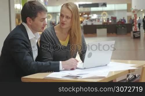 Two business people are sitting at table in trade centre and discussing their new enterprise lively. Laptop is standing in front of them.