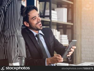 Two business people working together in office. Businessman sitting in office desk and using tablet while business woman standing next to him.