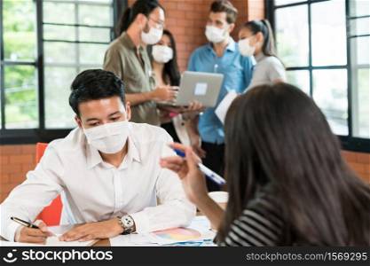 Two business person brainstorm in meeting room with group of interracial business worker team working in background. They wear face mask in new normal office prevent coronavirus COVID-19 spreading