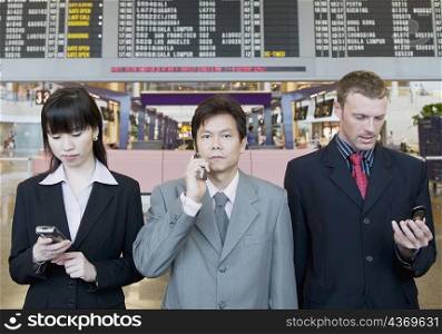 Two businessmen and a businesswoman holding mobile phones and standing at an airport