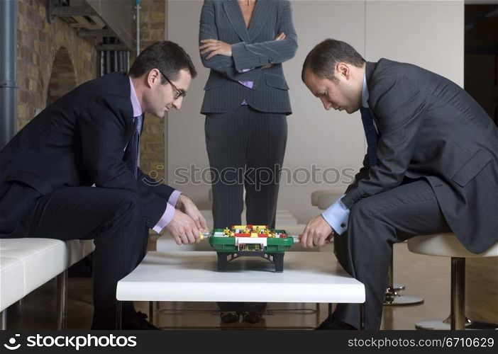 Two businessmen playing foosball with a businesswoman standing beside them