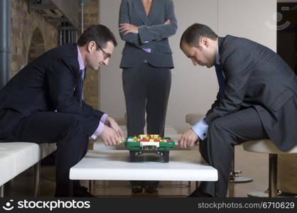 Two businessmen playing foosball with a businesswoman standing beside them