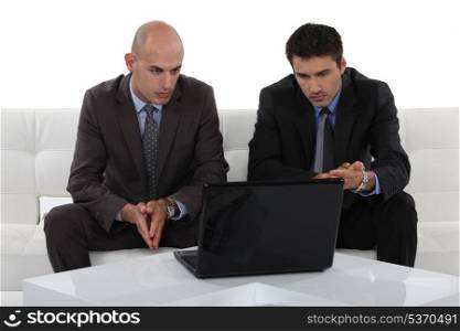 two businessmen sitting on a sofa and watching something on a laptop