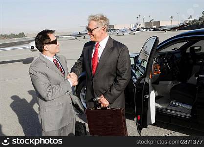 Two businessmen standing on landing strip in front of car and shaking hands.