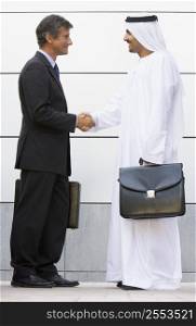 Two businessmen standing outdoors with briefcases shaking hands and smiling