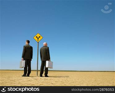 Two businessmen with briefcases standing by road sign in desert full length back view