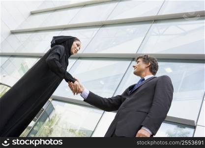 Two businesspeople standing outdoors by building shaking hands and smiling (selective focus)