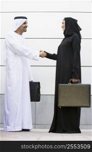 Two businesspeople standing outdoors with briefcases shaking hands and smiling