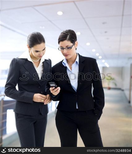 Two businesswomen looking at mobile phone screen on the office corridor.