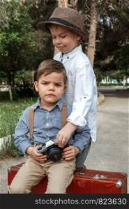 two Caucasian boy brothers in vintage clothes. One is sitting on an old suitcase, the other is trying to take his retro camera
