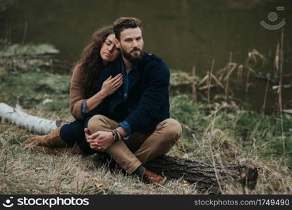 two caucasian lovers are sitting on the shore of the lake. Young couple is hugging on autumn day outdoors. A bearded man and curly woman in love. Valentine’s Day. Concept of love and family. two caucasian lovers are sitting on the shore of the lake. Young couple is hugging on autumn day outdoors. A bearded man and curly woman in love. Valentine’s Day. Concept of love and family.