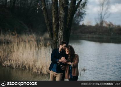 two caucasian lovers are standing near tree by the lake. Young couple is hugging on autumn day outdoors. A bearded man and curly woman in love. Valentine’s Day. Concept of love. two caucasian lovers are standing near tree by the lake. Young couple is hugging on autumn day outdoors. A bearded man and curly woman in love. Valentine’s Day. Concept of love.