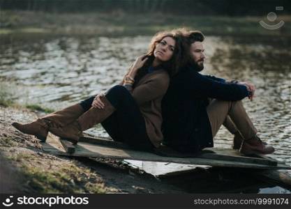 two caucasian lovers sitting on the pier by the lake. Young couple is hugging on autumn day outdoors. A bearded man and curly woman in love. Valentine’s Day. Concept of love and family. two caucasian lovers sitting on the pier by the lake. Young couple is hugging on autumn day outdoors. A bearded man and curly woman in love. Valentine’s Day. Concept of love and family.