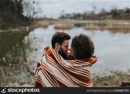 two caucasian lovers wrapped in blanket by the lake. Young couple is hugging on autumn day outdoors. A bearded man and curly woman in love. Valentine’s Day. Concept of love and family. two caucasian lovers wrapped in blanket by the lake. Young couple is hugging on autumn day outdoors. A bearded man and curly woman in love. Valentine’s Day. Concept of love and family.