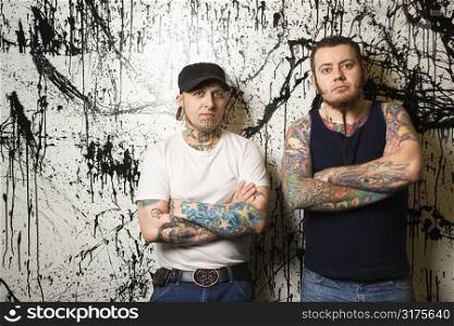 Two Caucasian tattooed men standing against paint splattered background.