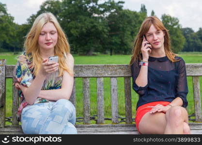 Two caucasian teenage girlfriends sitting on wooden bench in park calling with mobile phone