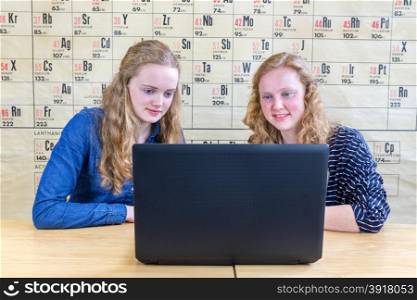 Two caucasian teenage girls looking at laptop in chemistry class in front of wallchart showing periodic table