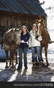 Two Caucasian women holding horses smiling at viewer with stable in background.
