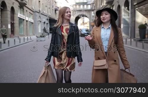Two charming multiracial girlfriends carrying shopping bags while walking along cobblestone street after good day shopping. Stunning young asian woman and her charming caucasian female friend chatting while shopping in boutique stores. Slow motion.