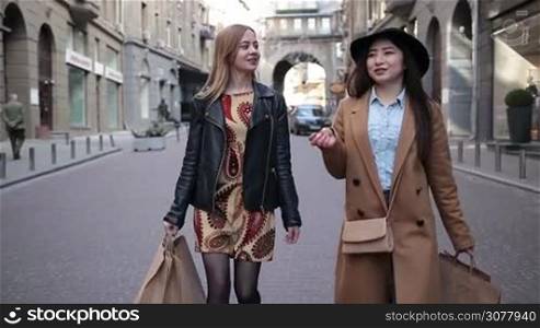 Two charming multiracial girlfriends carrying shopping bags while walking along cobblestone street after good day shopping. Stunning young asian woman and her charming caucasian female friend chatting while shopping in boutique stores. Slow motion.