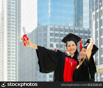 Two cheerful graduated woman wearing mortarboard and academic dress, holding diploma. Smiling women hugging each other on graduation ceremony and standing outdoors with city buildings background