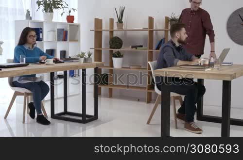 Two cheerful hipster freelancers discussing new startup business using laptop pc in modern office while asian female colleague browsing the net with digital tablet. Businesspeople working in office.