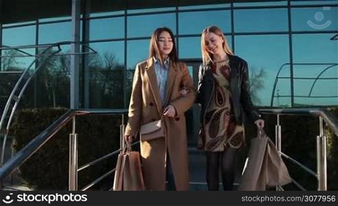 Two cheerful women friends coming back from shopping, enjoying a relaxed day together. Excited beautiful multiracial females walking out the shopping mall with purchases in packages and bags in the glow of amazing sunset. Slow motion.