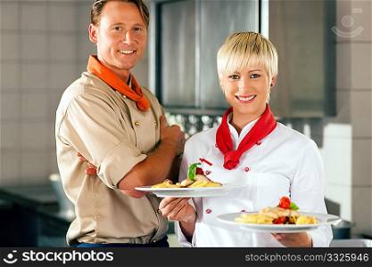 Two chefs in teamwork - man and woman - in a restaurant or hotel kitchen cooking delicious food, both are presenting the finished dishes